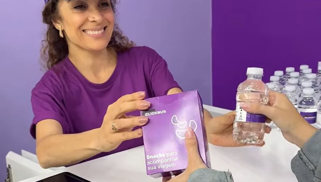 Mulher sorridente em estande roxo entrega um pacote de snacks ClickBus e uma garrafa de água a outra pessoa.