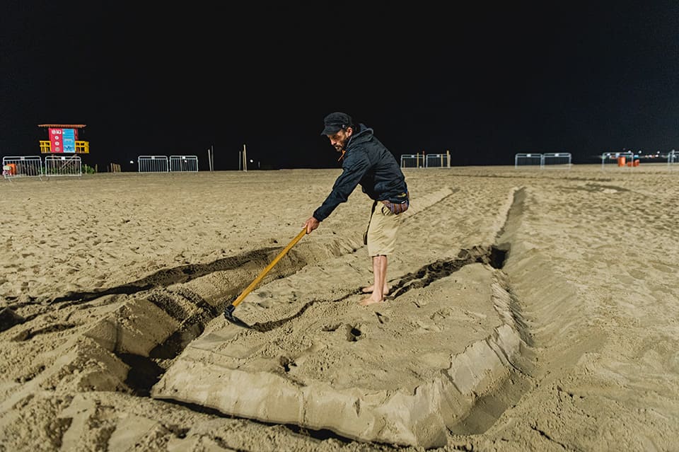 Homem trabalhando descalço na areia da praia durante a noite, usando uma enxada para escavar ou moldar uma grande forma na areia.