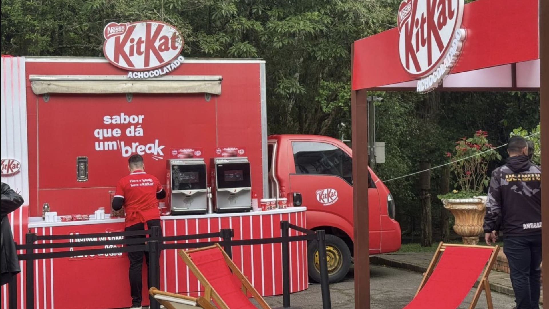 Ativação promocional da KitKat com um food truck vermelho e branco, onde um funcionário serve bebidas. Há espreguiçadeiras vermelhas e um toldo grande da marca.