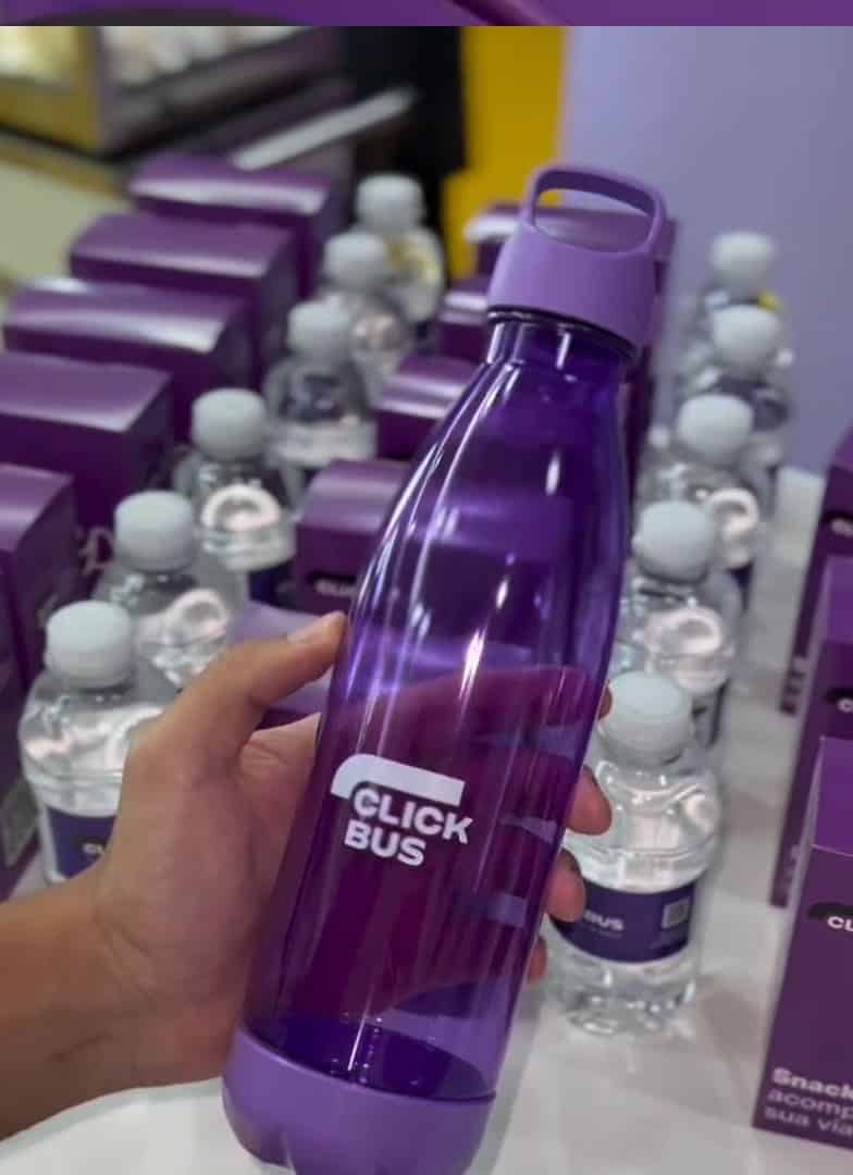 Uma mão segura uma squeeze roxa translúcida da ClickBus com fundo desfocado de várias garrafas de água e caixas de snacks roxos.