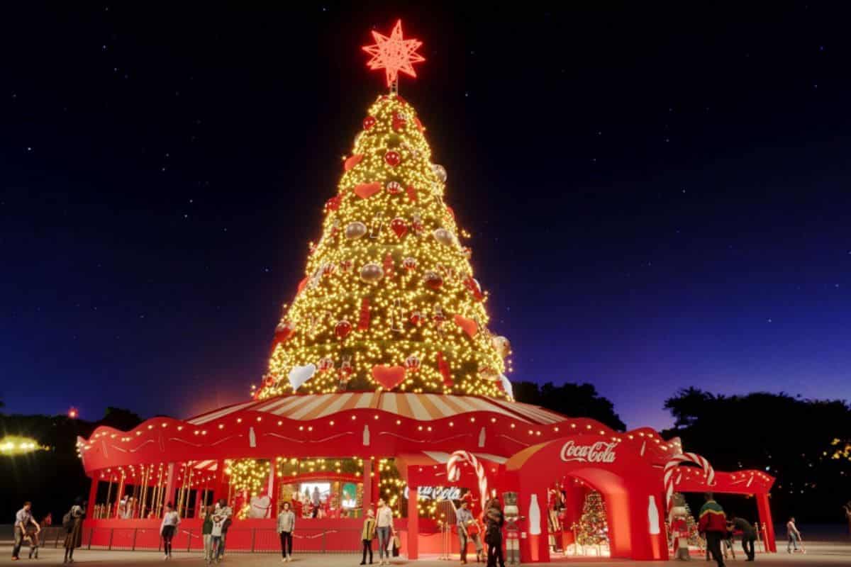 Papai Noel da Coca-Cola em frente a uma grande árvore de Natal iluminada e um carrossel.