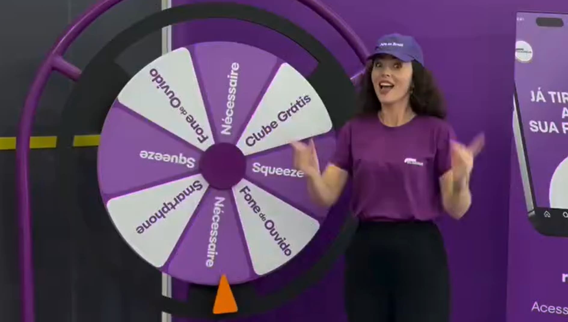 Mulher animada em camiseta roxa e boné, em pé ao lado de uma Roda da Fortuna roxa e branca com prêmios como "Smartphone", "Fone de Ouvido", "Squeeze" e "Clube Grátis".