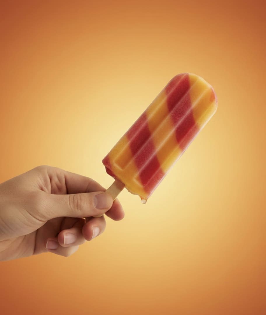 Mão segura picolé listrado de laranja e vermelho derretendo, contra um fundo laranja gradiente.