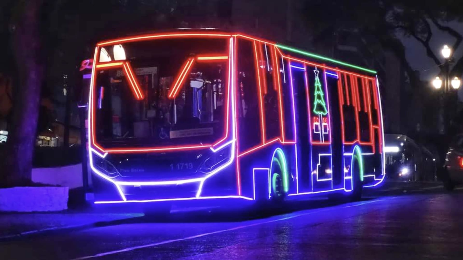 Ônibus urbano moderno completamente contornado por luzes LED de Natal nas cores azul, vermelho e verde, com um desenho de árvore de Natal verde no meio. O ônibus está na rua à noite.