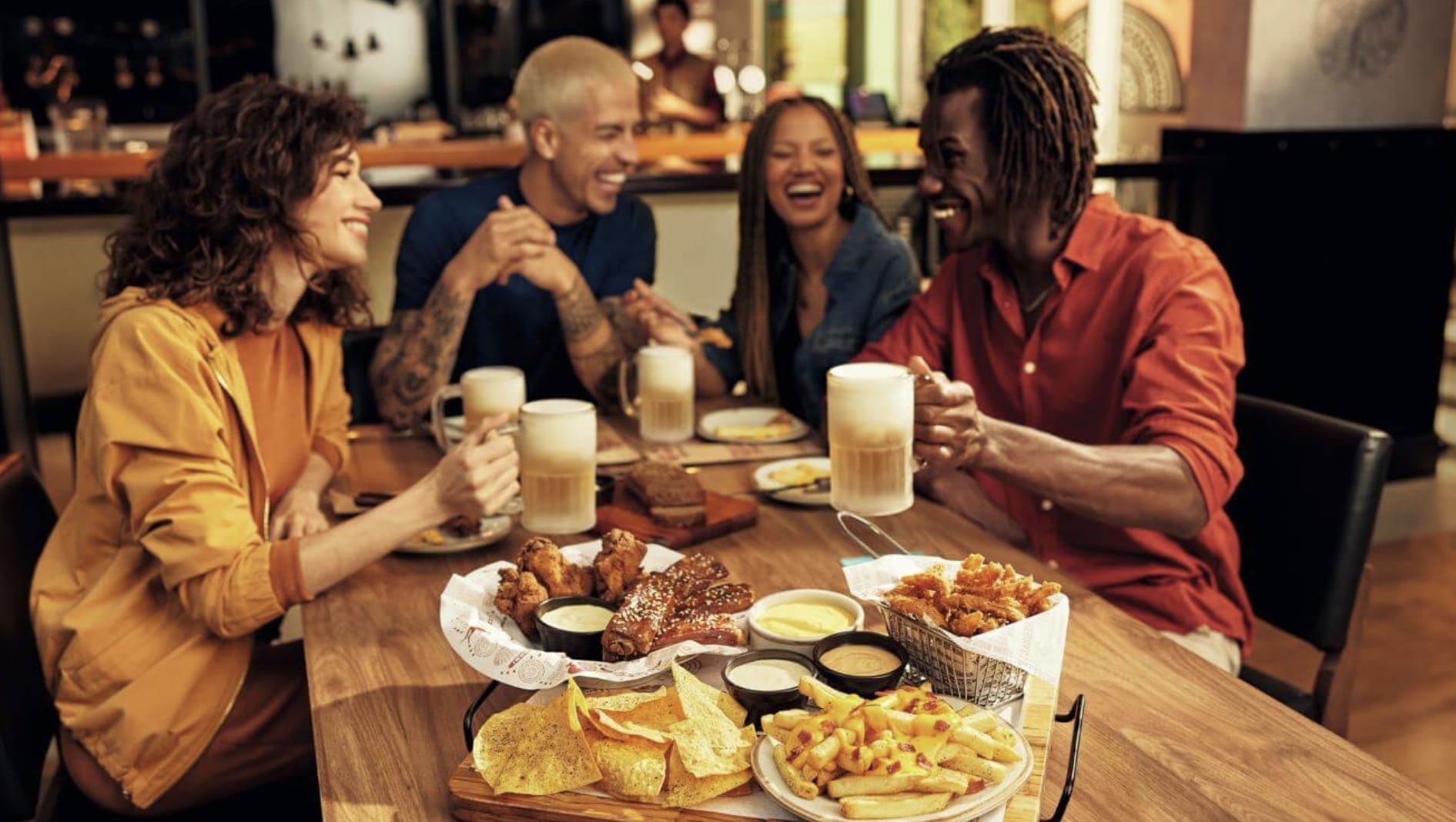 Quatro amigos rindo e brindando com canecas de chopp enquanto desfrutam de vários aperitivos na mesa do Outback.