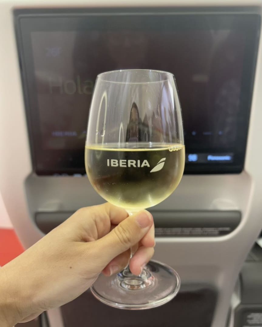 Mão segurando uma taça de vinho branco com o logo Iberia a bordo de um avião, com tela de entretenimento ao fundo.