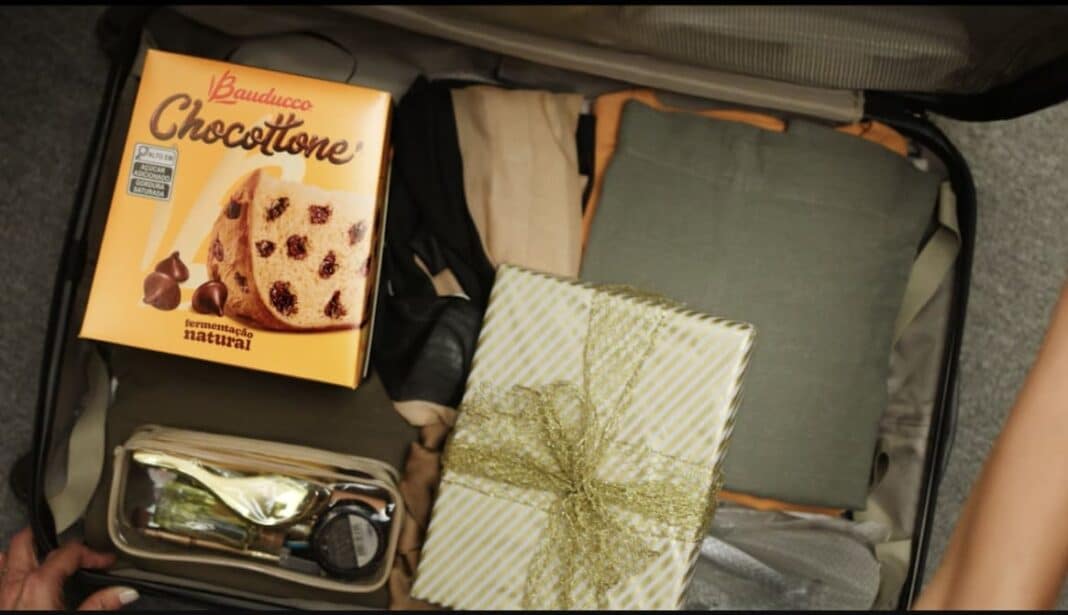 Uma mala de viagem aberta vista de cima, contendo roupas dobradas, um presente embrulhado em papel dourado e uma caixa amarela de Chocottone Bauducco.