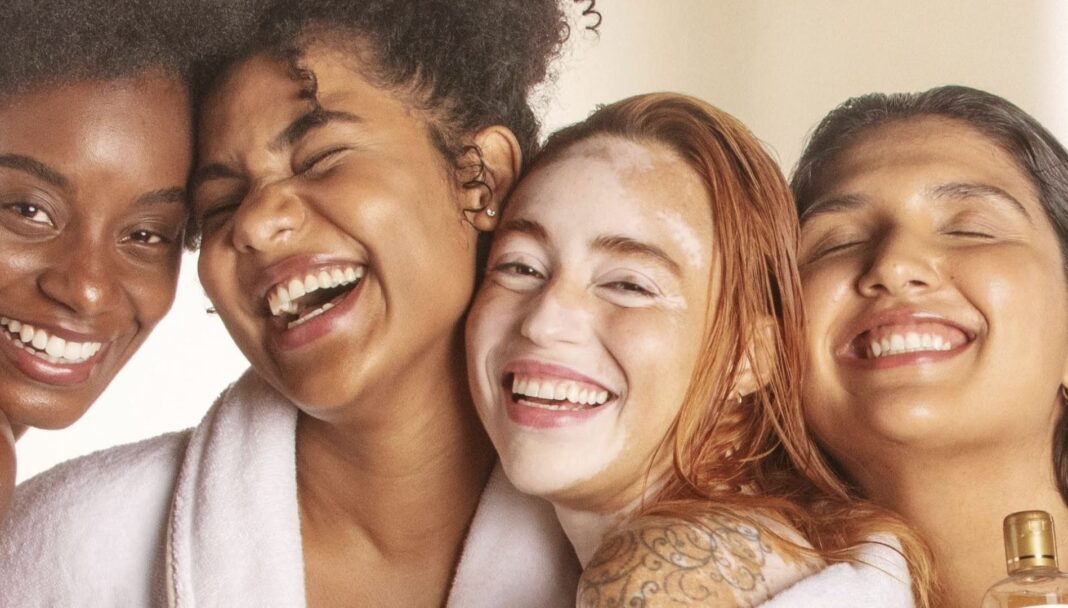 Quatro jovens adultas, de diversas etnias e tipos de pele, sorrindo e abraçadas.