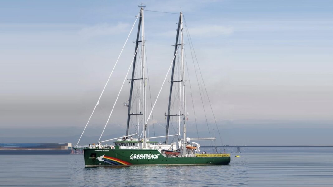 O navio Rainbow Warrior do Greenpeace navegando em águas calmas no Rio de Janeiro. O navio é verde com o logo da organização e um arco-íris na proa. Ao fundo, vê-se a Ponte Rio-Niterói sob um céu claro.