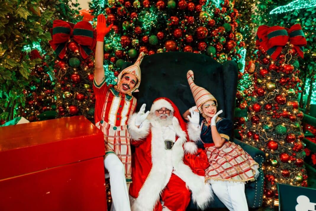 Papai Noel sentado em um trono de veludo verde, ladeado por dois ajudantes (elfos) sorridentes e caracterizados com roupas xadrez e chapéus pontudos. O cenário é ricamente decorado com árvores de Natal repletas de bolas vermelhas e douradas e luzes brilhantes ao fundo.
