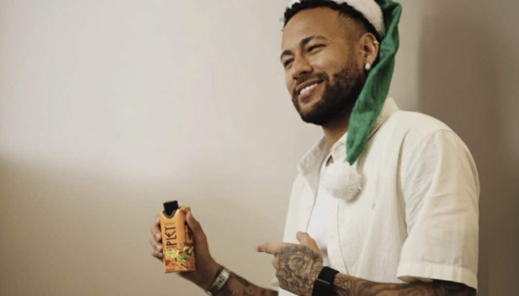 Neymar Jr. sorrindo, segurando uma caixinha de suco laranja/preto, usando um chapéu de Papai Noel verde e branco.