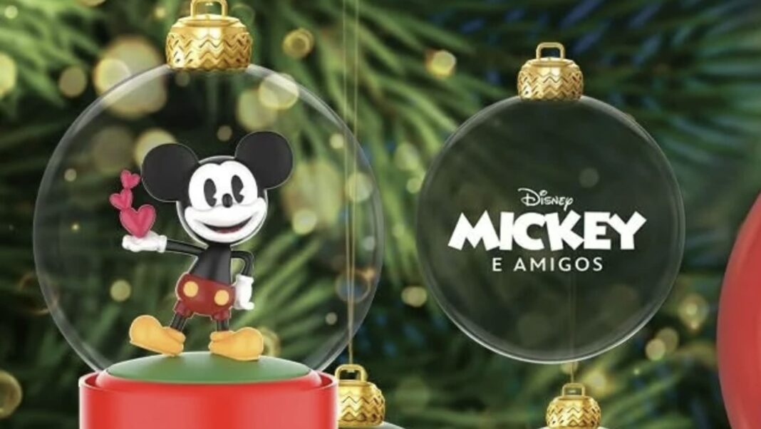 Miniatura do Mickey Mouse segurando corações dentro de uma bola de Natal transparente, ao lado de outra bola com o logotipo 'Disney Mickey e Amigos', com fundo de árvore de Natal desfocado.