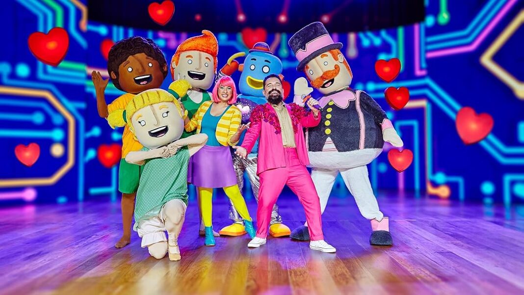 Personagens do Mundo Bita e dançarinos posam sorridentes em um palco colorido com temática digital e corações flutuantes.