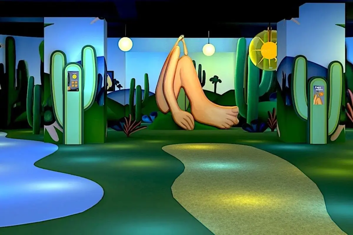 Cenografia de exposição de arte em ambiente escuro, com um grande corpo humano estilizado e pernas alongadas, em meio a grandes cactos verdes e paisagem de fundo azul.