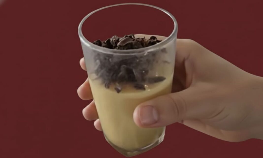 : Mão segurando um copo de vidro pequeno com um doce cremoso de cor amarelada, finalizado com raspas de chocolate amargo no topo. Fundo bordô sólido.