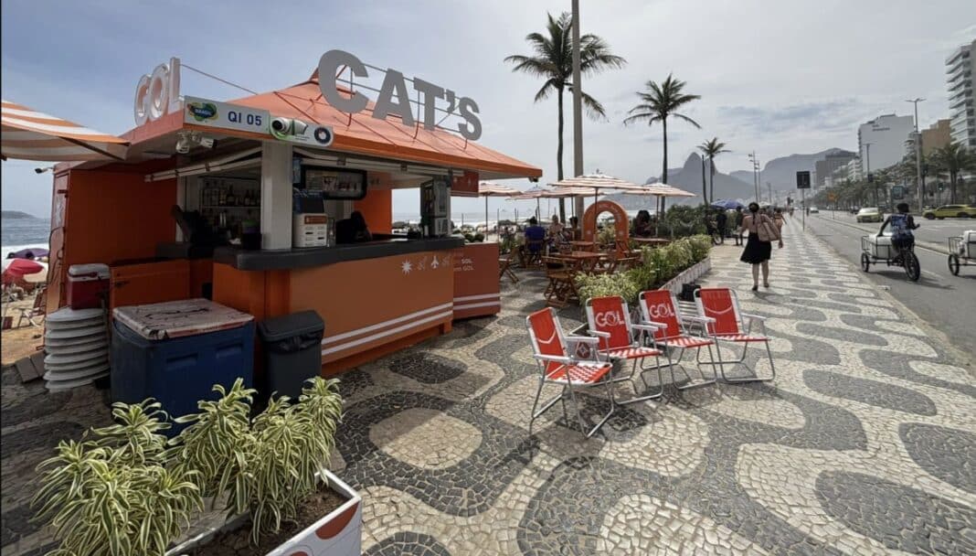 Quiosque laranja da GOL e CAT’S no calçadão de Ipanema, Rio de Janeiro, com cadeiras de praia alinhadas, coqueiros e o Morro Dois Irmãos ao fundo sob um céu claro.