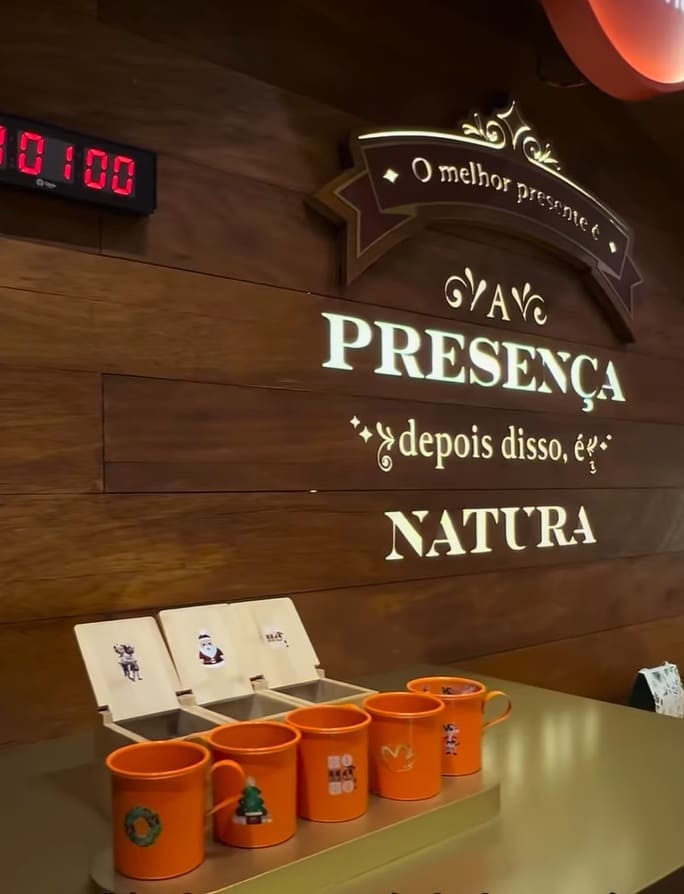 Painel de madeira em um evento da Natura com a frase em destaque: "O melhor presente é a PRESENÇA, depois disso, é NATURA". Abaixo, cinco canecas laranjas com temas natalinos e tampas com ilustrações aguardam retirada em uma bancada dourada. No canto superior esquerdo, um contador digital vermelho exibe o número "00100".