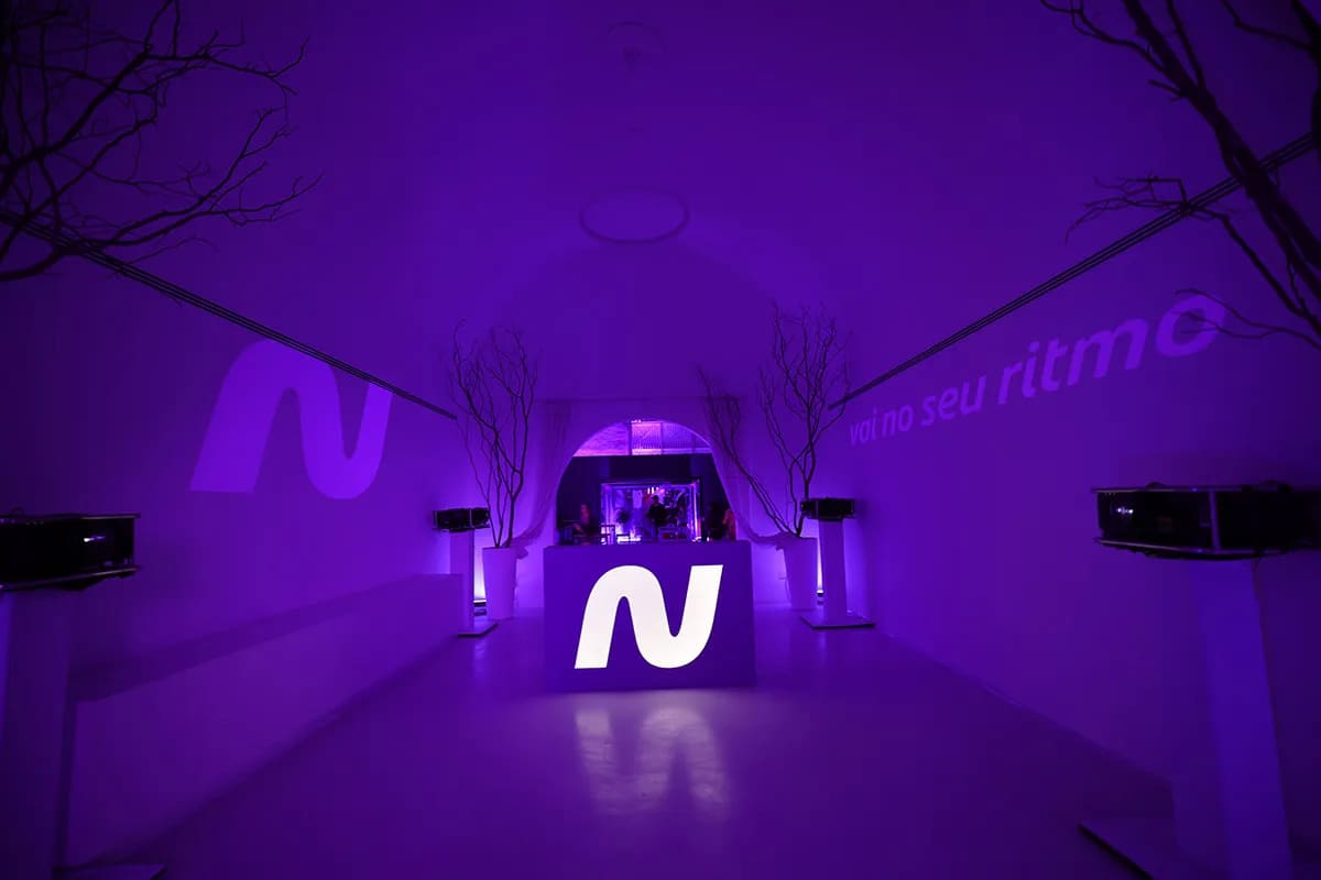 Corredor de um evento iluminado com luz roxa vibrante, apresentando o logotipo "N" da Netshoes projetado nas paredes e em um balcão central iluminado ao fundo.