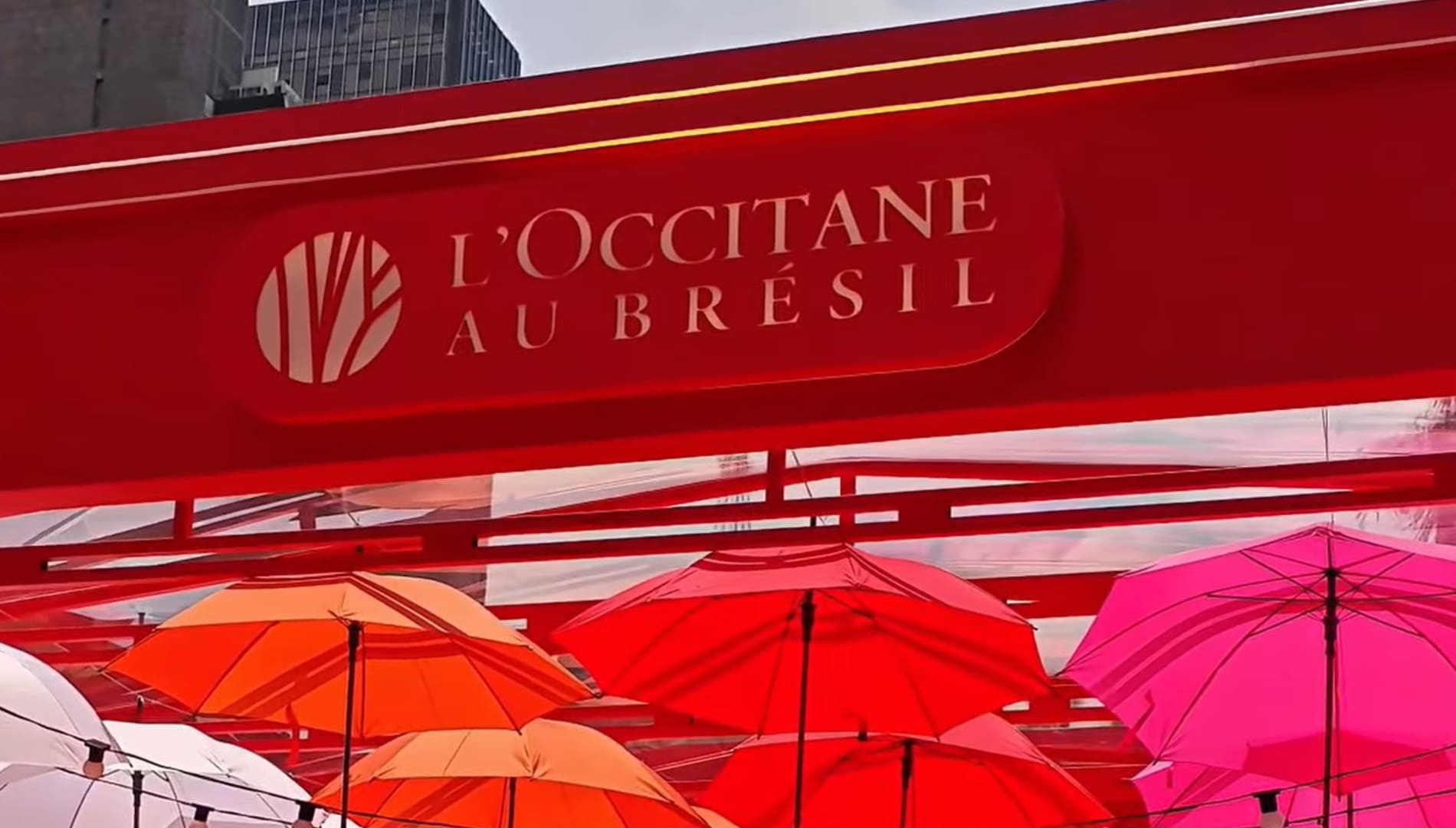 Banner vermelho da L'Occitane Au Brésil sobre guarda-chuvas coloridos.