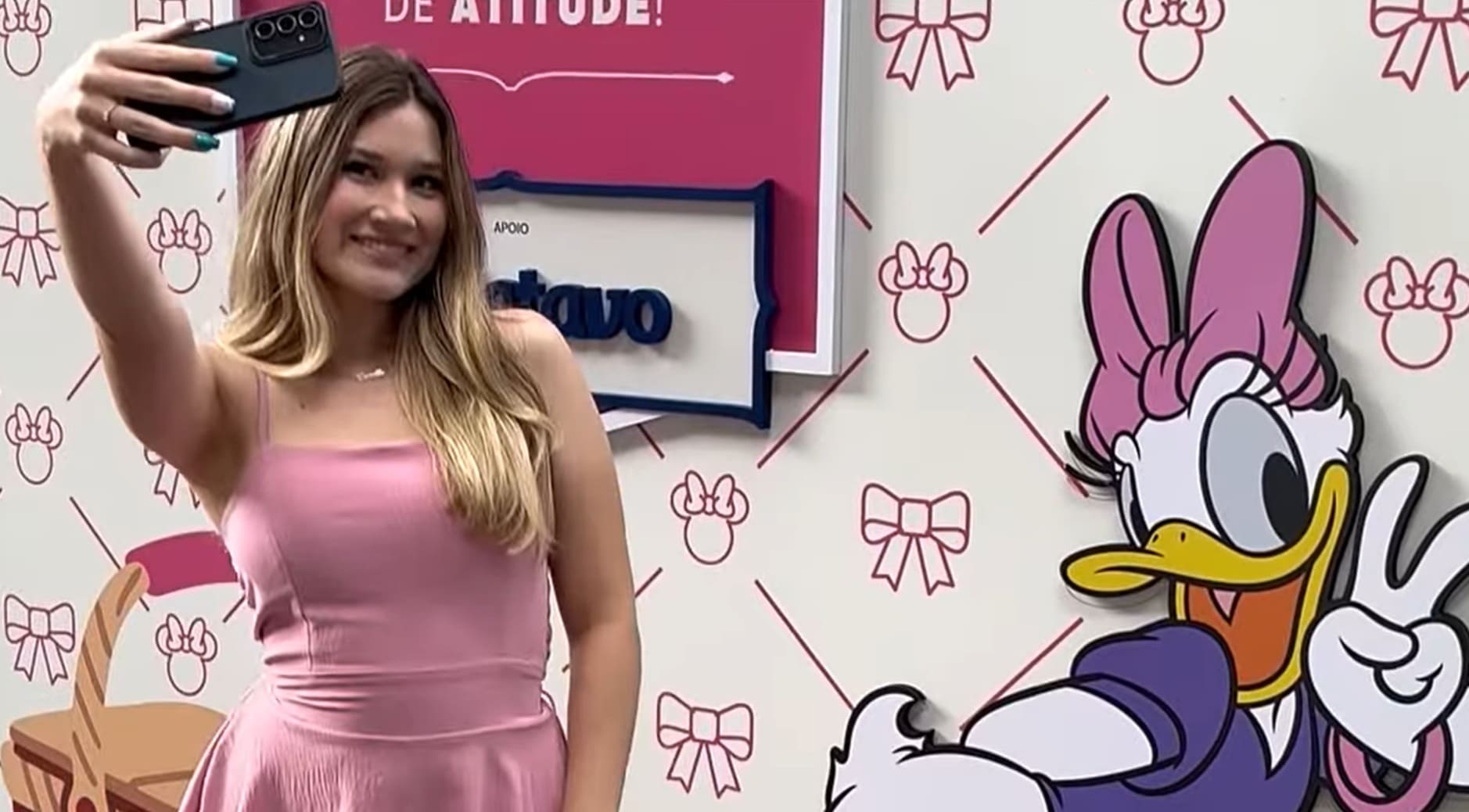 Mulher sorrindo, vestindo rosa, tirando uma selfie em frente a um painel temático com a Margarida da Disney e o apoio da marca Batavo.