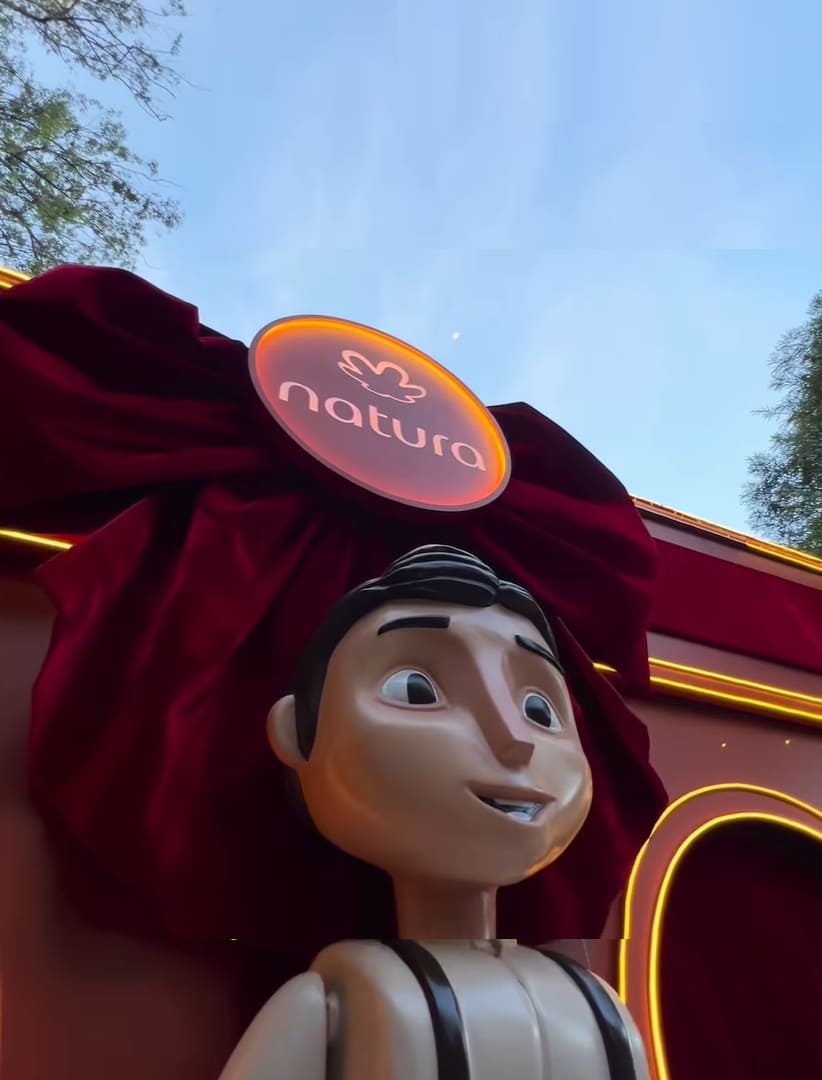 Boneco animatrônico ou manequim de um rapaz de pele clara e cabelo escuro, vestido com um uniforme de jardineiro ou zelador, posicionado à frente de uma decoração natalina vermelha com um grande laço de veludo e o logotipo da Natura brilhante.