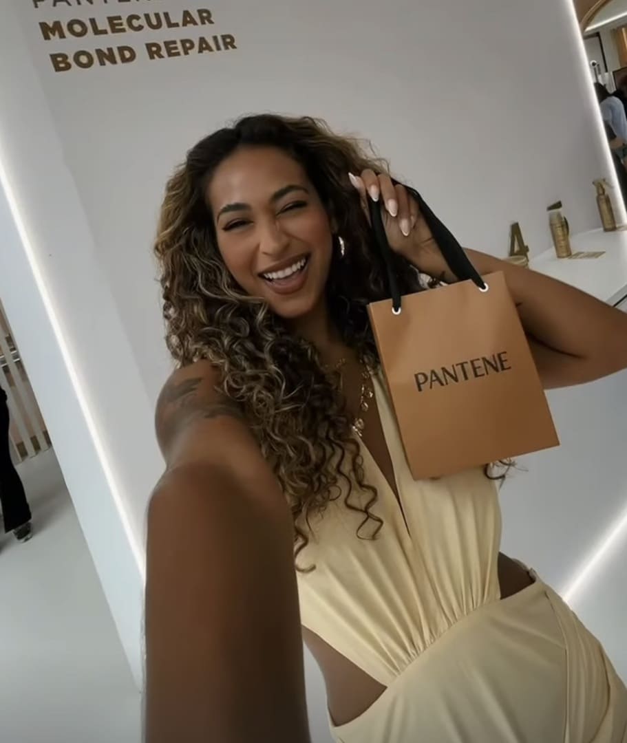 Mulher sorridente com cabelos cacheados e vestido amarelo tirando uma selfie, segurando uma pequena sacola de brinde da Pantene.