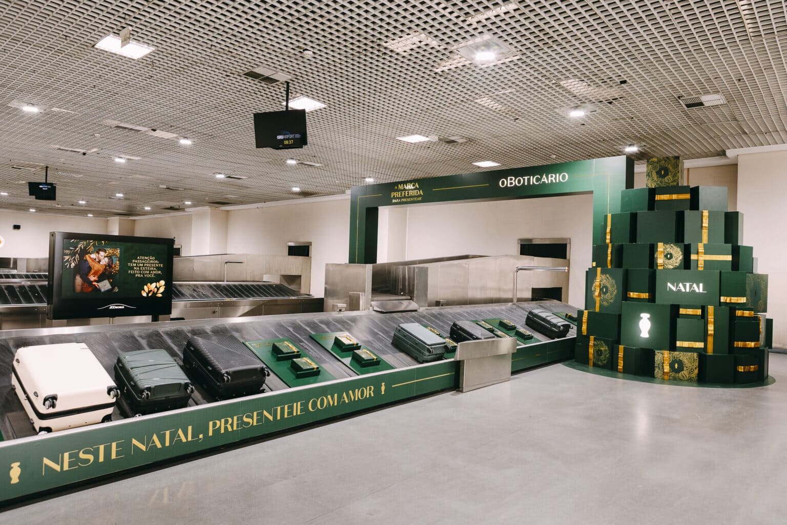 Cenário natalino da O Boticário em uma área de desembarque de aeroporto. Uma pilha de caixas de presente verdes e douradas em formato de árvore está ao lado de uma esteira de bagagens que exibe malas e caixas menores de presentes da marca, com mensagens de Natal.