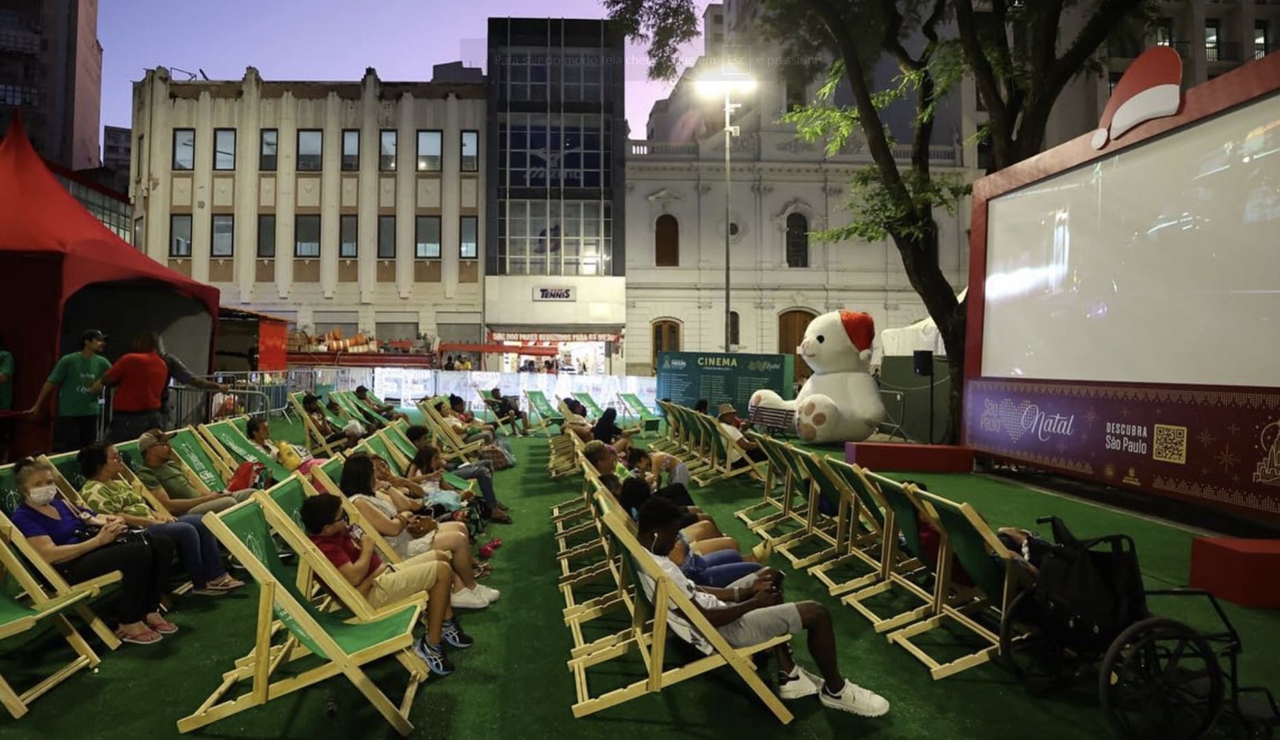 Público assiste a uma sessão de cinema de Natal ao ar livre em uma praça urbana, sentados em longas filas de cadeiras de praia verdes sobre um gramado, com um grande urso de pelúcia decorativo ao lado da tela.