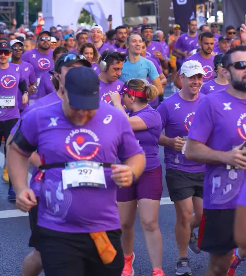 Multidão de corredores participantes da 99ª Corrida de São Silvestre usando camisetas roxas oficiais em uma via urbana.