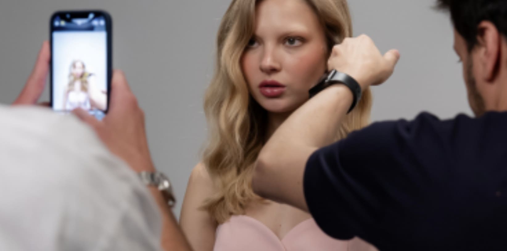 Uma modelo loira está sendo fotografada em um estúdio, com uma pessoa ajustando seu cabelo enquanto outra tira uma foto com um smartphone.