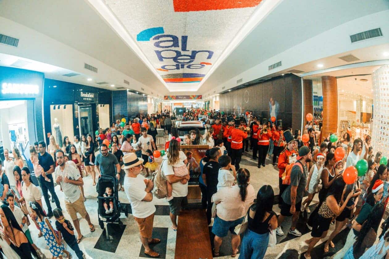 Vista ampla do corredor de um shopping center lotado de pessoas durante um evento de Natal. À direita, uma banda marcial vestindo uniformes vermelhos clássicos toca instrumentos de sopro. Famílias com crianças, carrinhos de bebê e balões coloridos preenchem o espaço. No teto, destaca-se um grande letreiro luminoso com a palavra "Alpendre".