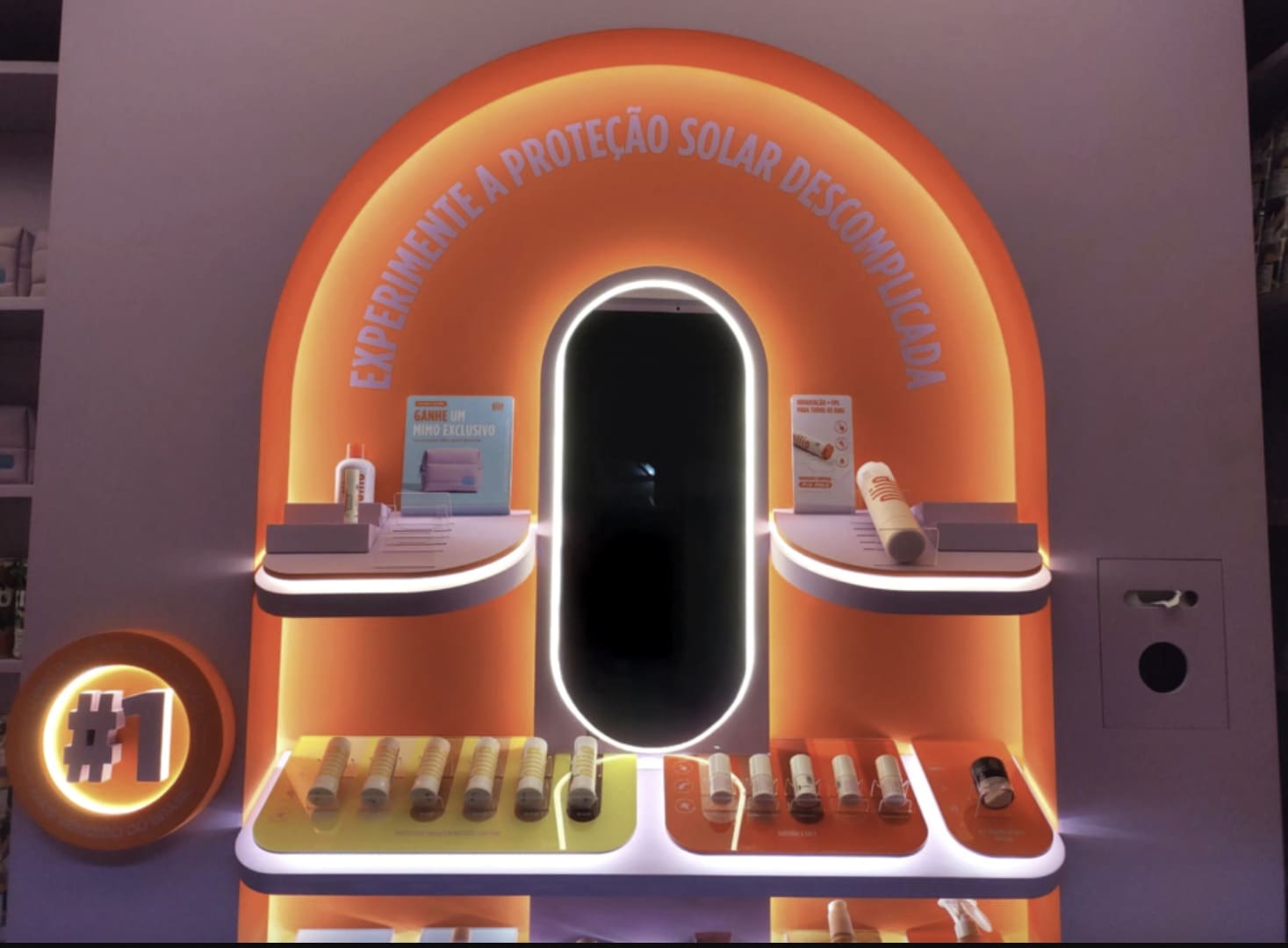 Close de um display expositor da Ollie com iluminação em LED laranja e lilás. O display possui um espelho central oval cercado pela frase "Experimente a proteção solar descomplicada", com prateleiras contendo diversos protetores solares em bastão e sinalizações promocionais.