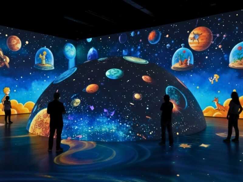 Sala de exposição imersiva com projeções em 360 graus do universo de O Pequeno Príncipe, apresentando uma grande cúpula central e paredes cobertas por ilustrações de planetas e estrelas.