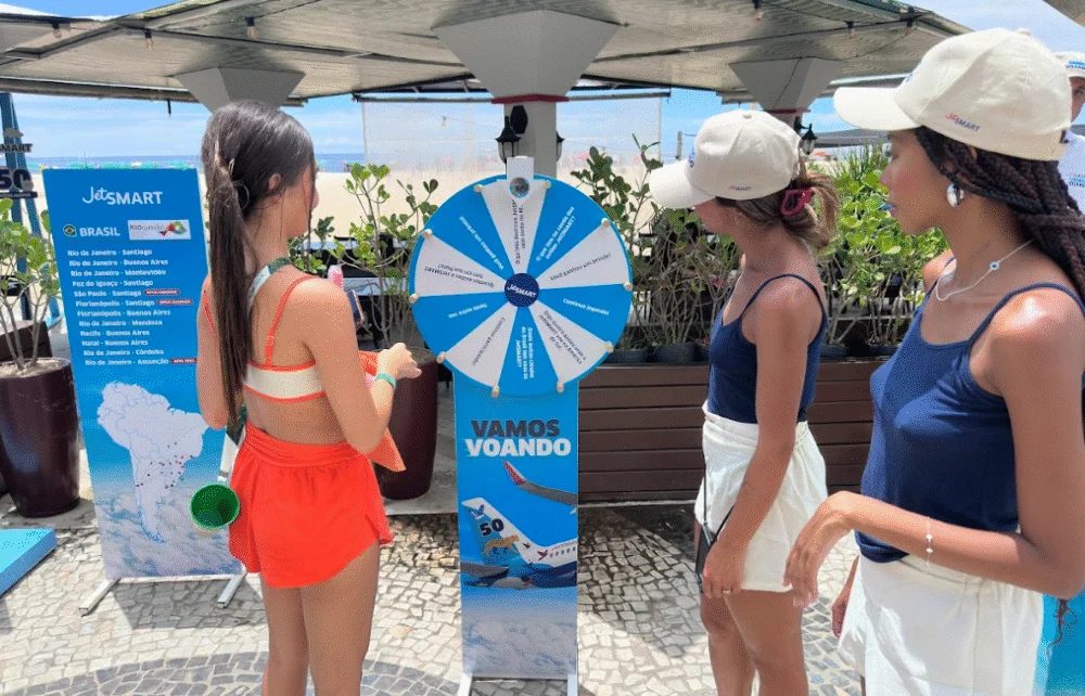 Três jovens participam de uma ativação da JetSMART em um quiosque na praia, interagindo com uma roleta de prêmios azul e branca com o slogan "VAMOS VOANDO".