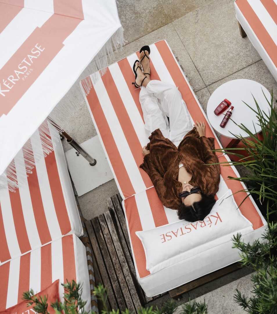 Vista aérea de uma pessoa descansando em uma espreguiçadeira com listras brancas e laranjas. Ao lado, há uma mesa redonda branca com produtos Kérastase da linha Soleil. Um guarda-sol com o logotipo da marca aparece no canto superior esquerdo. A pessoa usa óculos escuros, camisa marrom e calça branca.