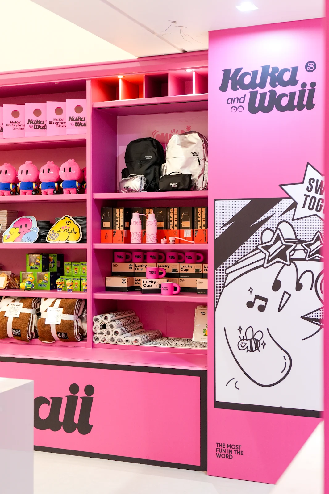 Prateleiras cor-de-rosa de uma loja Kwai com produtos temáticos: bonecos de porco rosa, mochilas, garrafas de água, canecas "Lucky Cup" e rolos de papel de embrulho ou tecido.