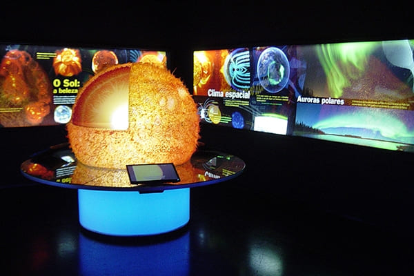 Interior do Museu Catavento exibindo uma maquete tridimensional e iluminada do Sol com um corte transversal que mostra suas camadas internas. Ao fundo, painéis digitais explicativos com os títulos "O Sol", "Clima espacial" e "Auroras polares".