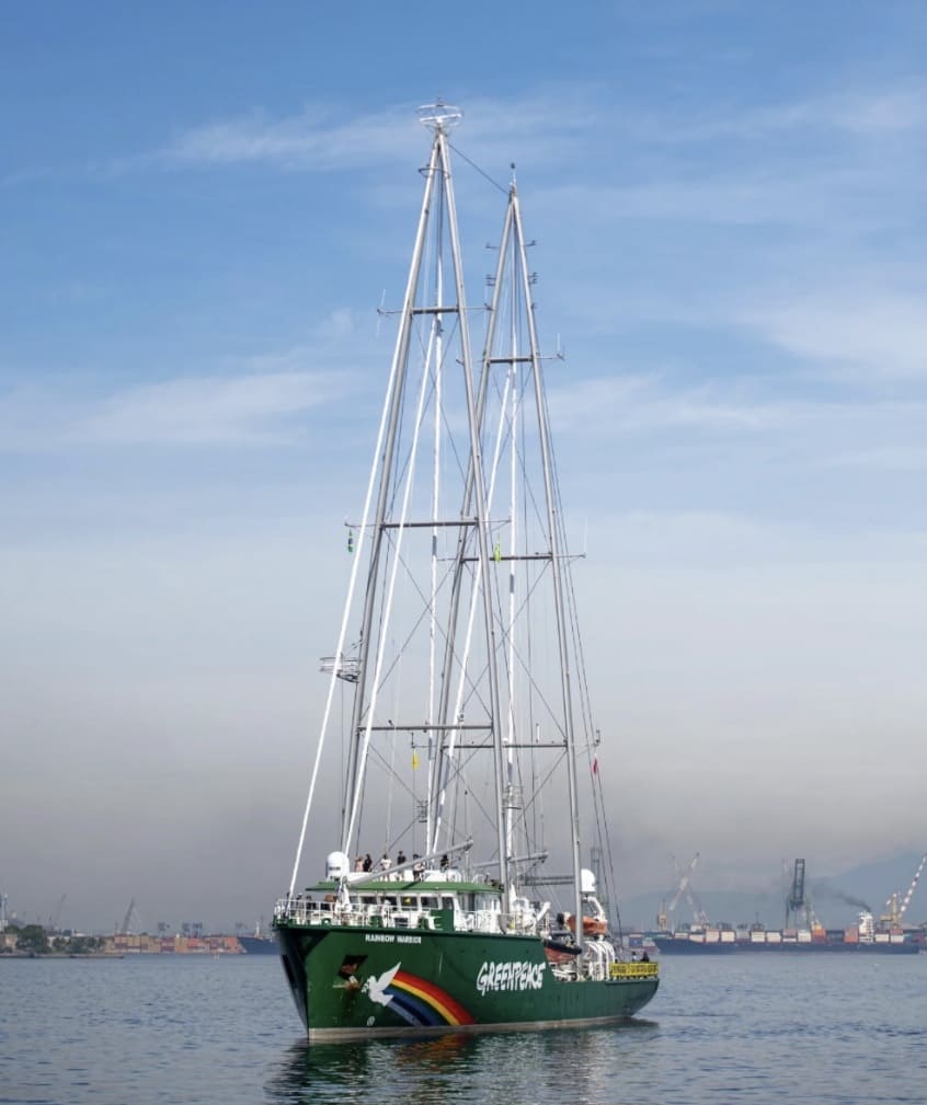 Fotografia do navio Rainbow Warrior do Greenpeace em um porto. A embarcação é verde com dois mastros prateados muito altos. Na proa, há uma pintura de um arco-íris e uma pomba branca. O céu está claro e, ao fundo, veem-se guindastes portuários e outros navios de carga.
