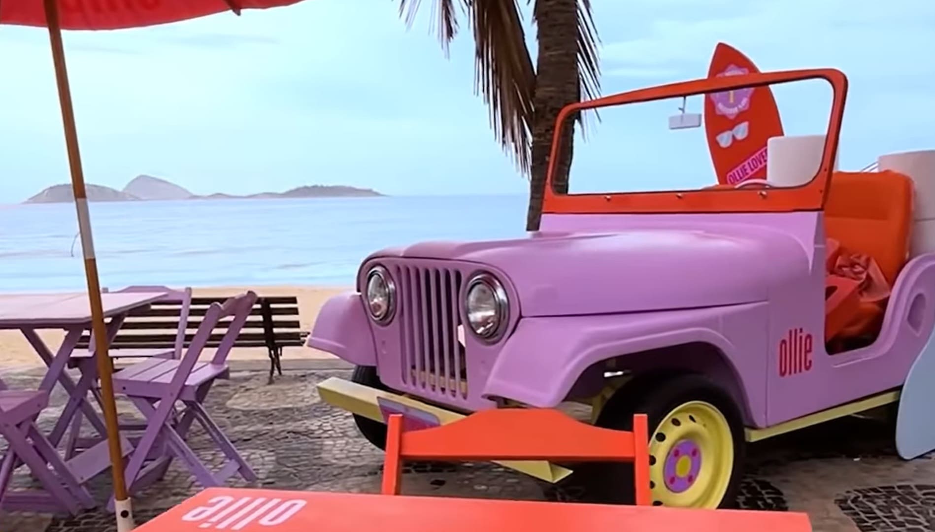Jipe lilás e laranja da marca Ollie, sem teto e com uma prancha de surfe, estacionado no calçadão de Ipanema, Rio de Janeiro, com vista para o mar e o Morro Dois Irmãos.