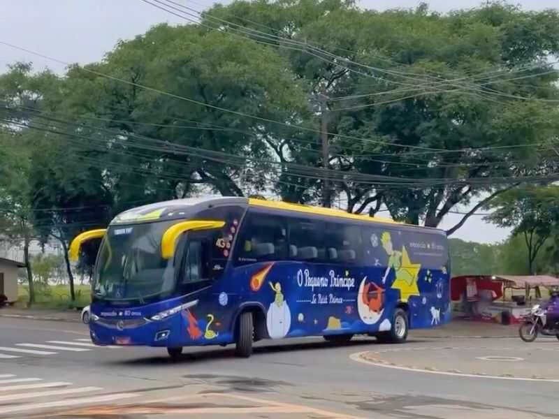 Ônibus de viagem azul decorado com ilustrações do livro "O Pequeno Príncipe", circulando em uma via urbana com árvores ao fundo.
