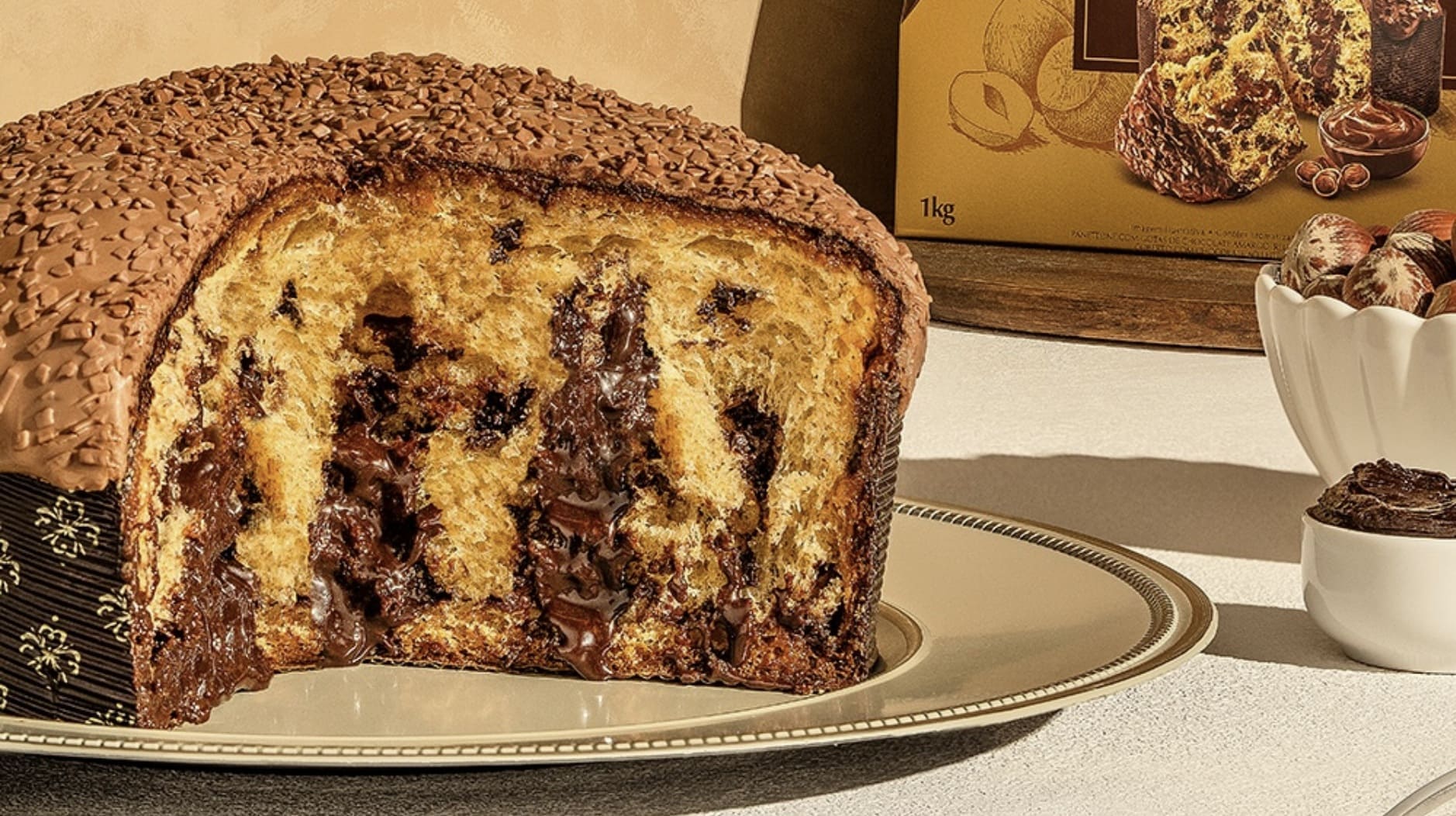 Fatia grossa de um panetone recheado com creme e gotas de chocolate derretido, coberto com uma camada de chocolate e granulado, servido em um prato com uma borda dourada.