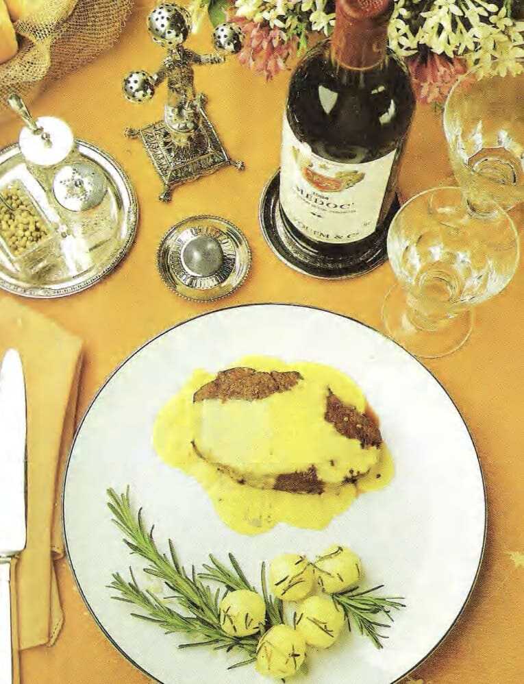 Prato de carne com molho amarelo acompanhado de batatas, alecrim e uma garrafa de vinho tinto sobre mesa amarela.