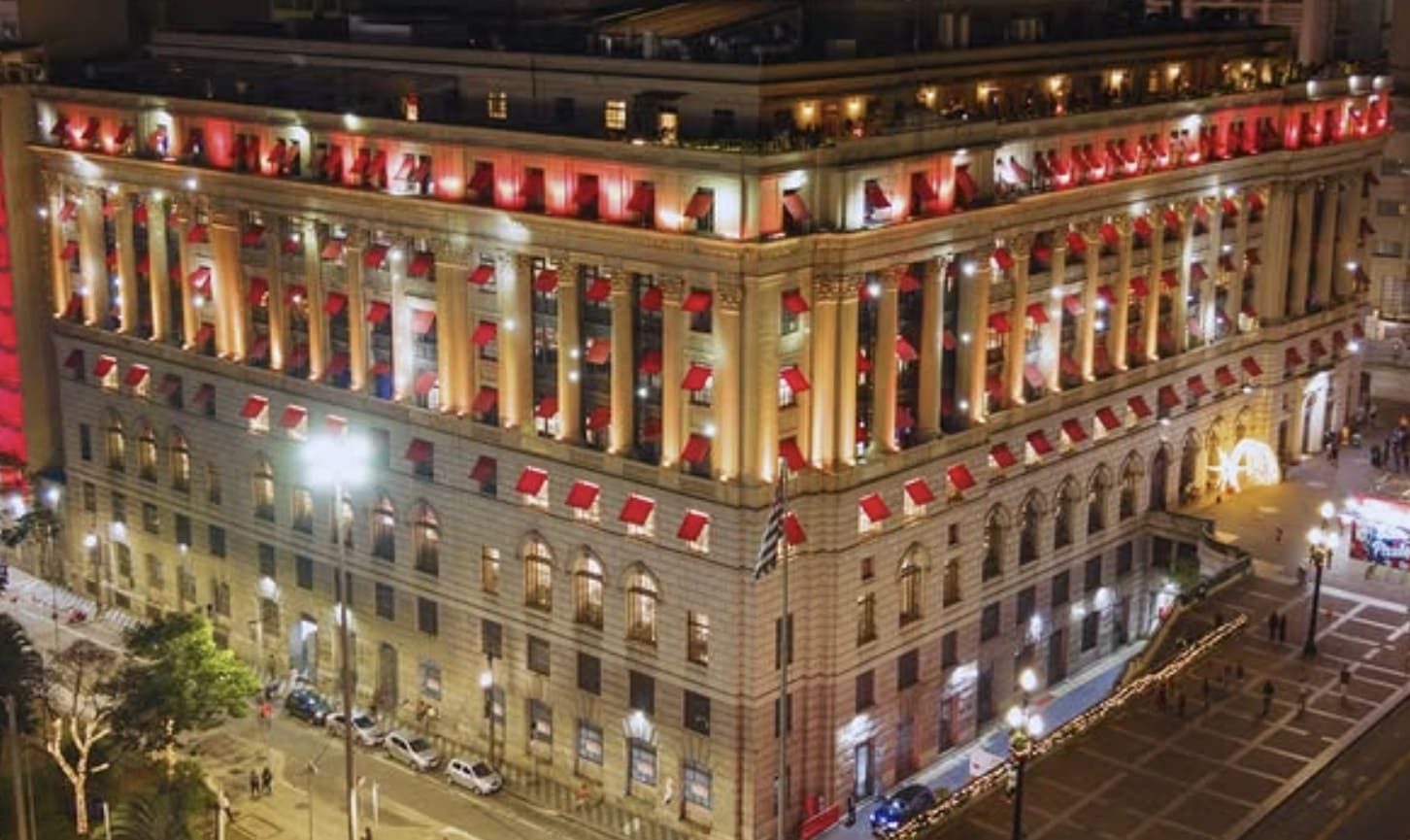 Edifício histórico e imponente, iluminado à noite com luzes quentes e detalhes em vermelho nas janelas, realçando sua arquitetura clássica.