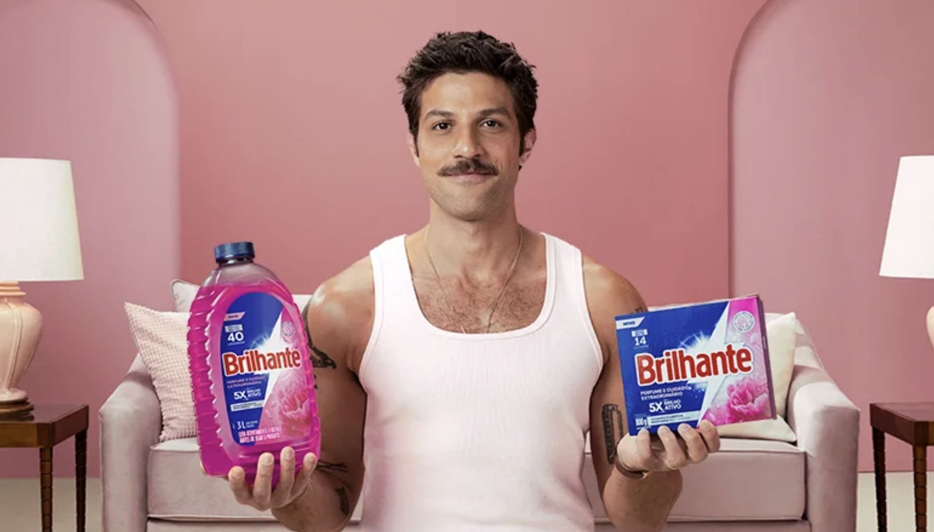 O ator Chay Suede sorri segurando dois produtos de limpeza da marca Brilhante (um sabão líquido e um em pó) em um cenário de sala com tons de rosa.