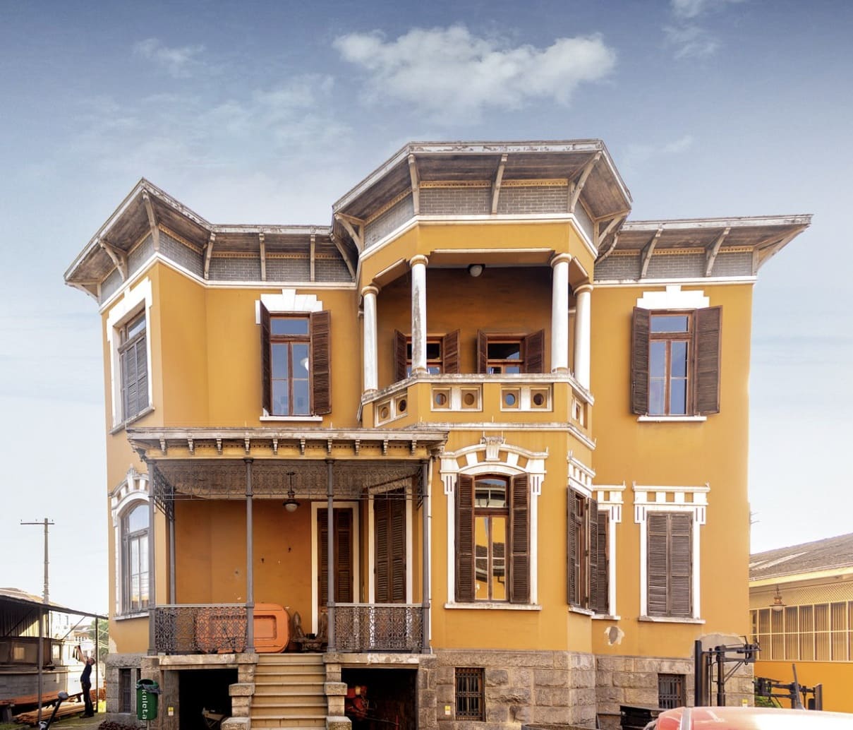 Fachada de um casarão histórico de dois andares em estilo eclético, pintado em amarelo ocre com detalhes brancos. O edifício possui varandas com colunas, janelas de madeira escura e telhado com beirais detalhados.