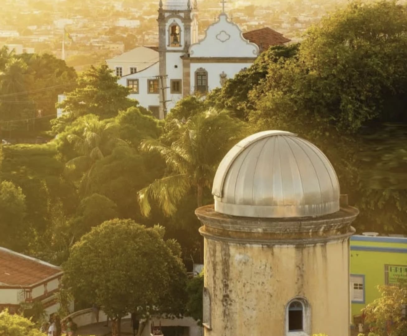 Fotografia aérea parcial do Alto da Sé em Olinda, mostrando em primeiro plano a cúpula prateada do Observatório Astronômico e, ao fundo, a fachada branca de uma igreja histórica cercada por vegetação densa sob uma luz dourada.