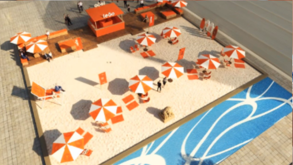 Vista aérea de uma ativação da marca Leão simulando uma praia urbana. O espaço retangular possui areia branca, diversos guarda-sóis listrados em laranja e branco, espreguiçadeiras e um quiosque laranja com o logo 'Leão'. Ao lado, há uma área azul que remete ao mar e um deck de madeira com assentos.