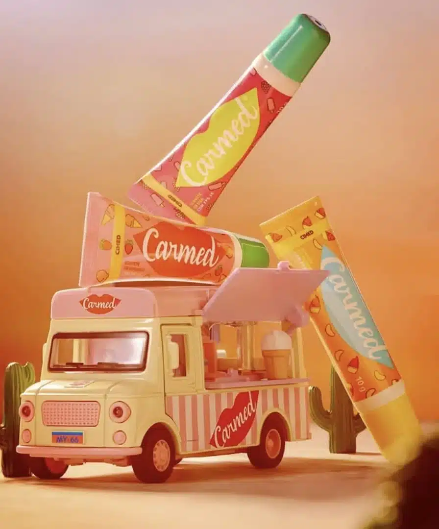 Um caminhão de sorvete de brinquedo decorado com o logotipo da Carmed, cercado por três tubos gigantes de hidratante labial Carmed Sorvetes em cores vibrantes (rosa, laranja e amarelo). O cenário tem tons quentes de pôr do sol e pequenos cactos ao fundo.