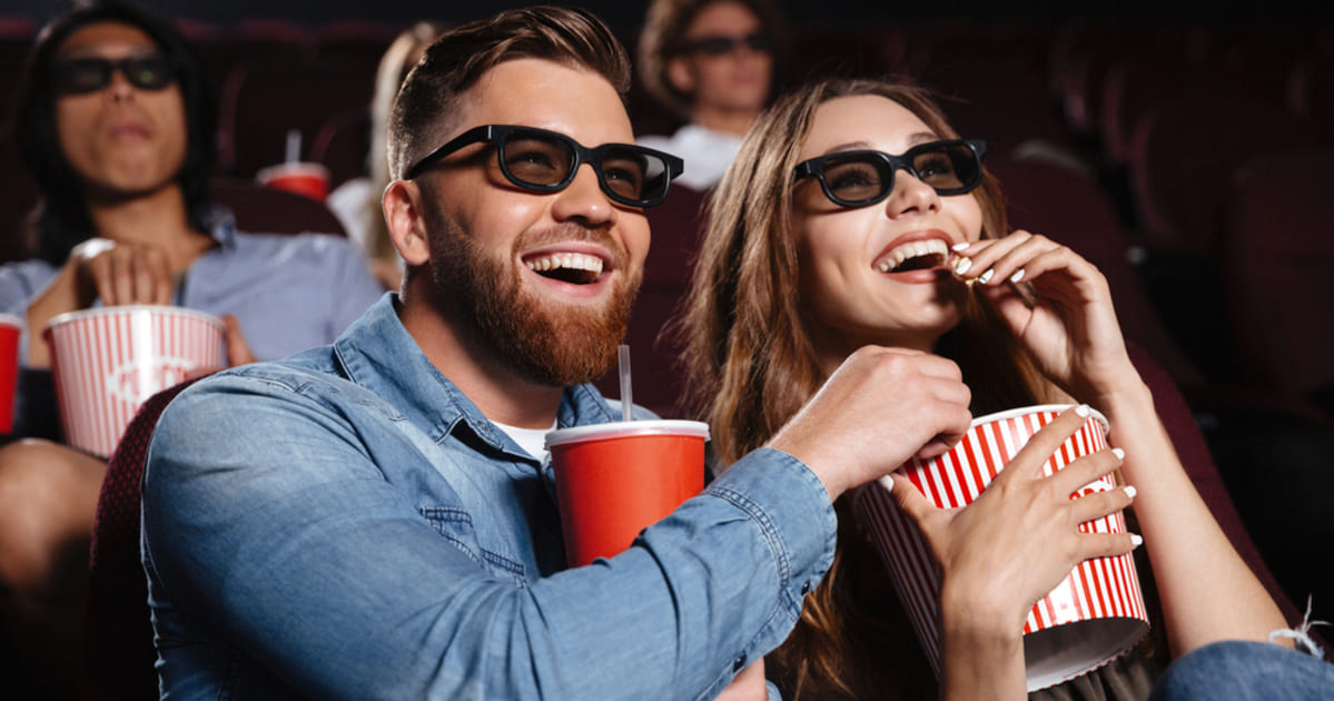 Casal jovem sorridente em uma sala de cinema, usando óculos 3D e comendo pipoca enquanto assiste a um filme.