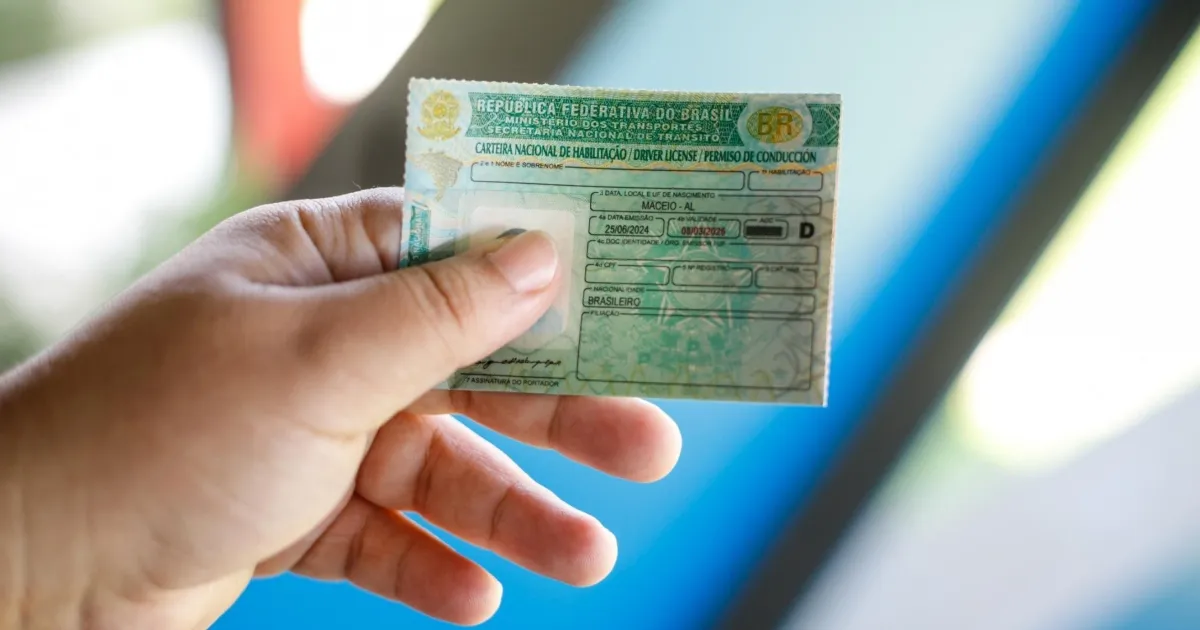 Uma mão segurando uma Carteira Nacional de Habilitação (CNH) brasileira física, com foco no documento. Ao fundo, um ambiente iluminado e desfocado.